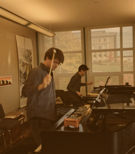 Photo of two pianists rehearsing at two pianos. Each is holding a mallet midair about to strike a woodblock on the piano desk.